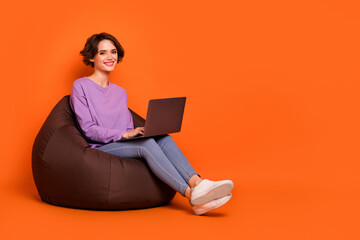 Full size portrait of pretty positive lady sitting cozy bag use wireless netbook isolated on orange color background