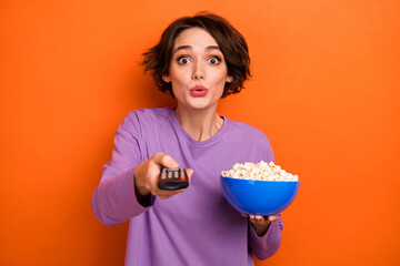Portrait of impressed nice girl hold popcorn plate switch tv program isolated on orange color background