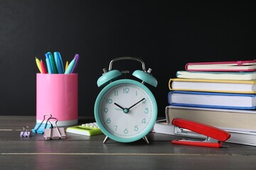 Alarm clock and different stationery on wooden table near blackboard. School time