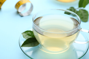 Glass cup of diet herbal tea and green leaves on light blue background, closeup