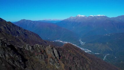 Stunning bird's eye view on the rosa khutor village drowning in the forested valley among majestic caucasian mountains. rocky slopes and mountain snowy peaks on the clear sky background.