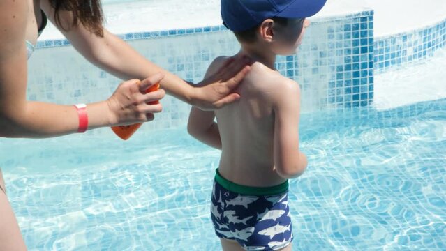 Mom applies sunscreen to the back of a 5-year-old kid standing by the pool.