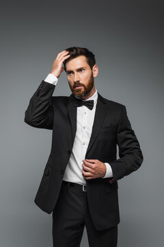 bearded man in black tuxedo with bow tie adjusting hair isolated on grey.