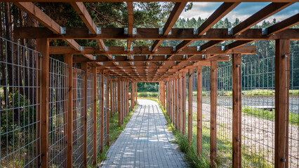 Wooden pergola (treillage) above cobblestone sidewalk leading along lake shoe and forest. One-point perspective.