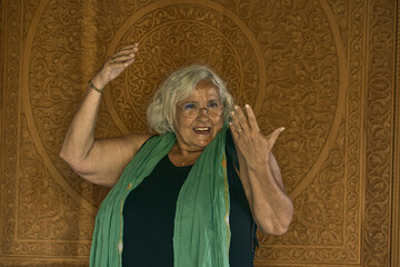Mujer senior contenta con vestido negro y pañuelo verde posando en una pared con decoración árabe