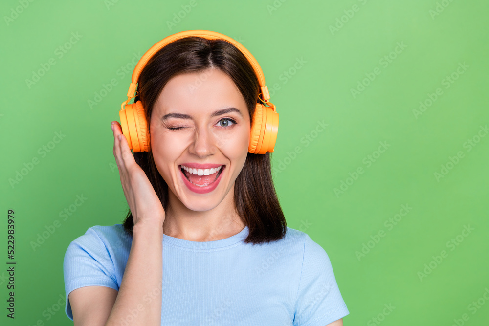 Poster portrait of adorable cheerful person arm touch headphones eye wink toothy smile isolated on green co