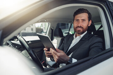 Handsome businessman working on digital tablet while sitting on driver seat in luxury car