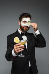 happy man in suit with clay mask on face holding cocktail isolated on grey.