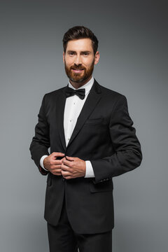 bearded man in black tuxedo with bow tie smiling isolated on grey.
