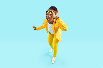 Fototapeta na wymiar Full body happy cheerful Afro American woman wearing stylish yellow suit and trendy glasses standing isolated on blue background, looking at camera, winking her eye and smiling. Party, fashion concept