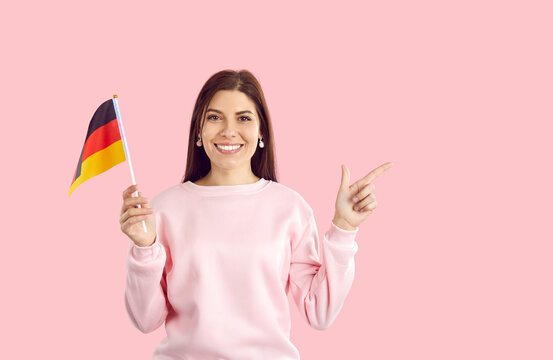 Happy cheerful beautiful young German woman in casual sweatshirt standing isolated on pastel pink background, holding flag of Germany, pointing her index finger at copy space on the right, and smiling