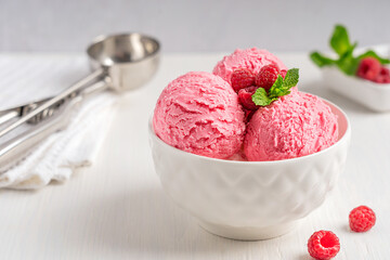 Homemade organic raspberry sorbet or gelato of pink color decorated with red berries and mint leaves served in bowl on white wooden table with ice cream scoop as sweet refreshing dessert or snack