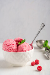 Scoops of cold homemade raspberry ice cream or sorbet of pink color decorated with red berries and mint leaves served in bowl with spoon on white wooden table as sweet refreshing dessert or snack