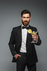 bearded man in elegant suit holding glass with cocktail and posing with hand in pocket isolated on grey.