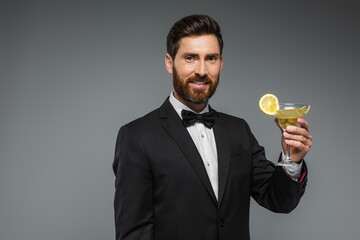 happy man in elegant suit with bow tie holding glass of cocktail isolated on grey.