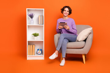 Full body photo of attractive satisfied lady sitting chair use tablet write message isolated on orange color background