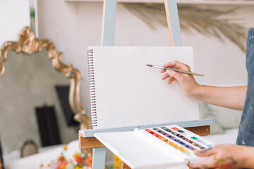 Closeup of student in apron painting in watercolor in an empty album on easel. Education, hobby, art concept.