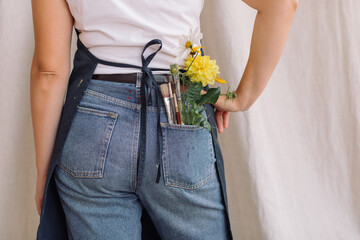 Flowers and brushes in the pocket of jeans of a young artist in apron. Back view. Creative. Authentic.