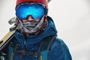 Equipped skier holding skis on his shoulder and looking directly at the camera, portrait. Man with winter equipment in the mountains at a ski resort - obrazy, fototapety, plakaty