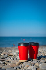 Fototapeta na wymiar Close-up of couple of paper coffee cups on pebble beach. Blue sea on background. Hot take away drinks.