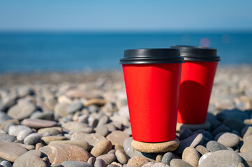 Close-up of couple of paper coffee cups on pebble beach. Blue sea on background. Hot take away drinks.
