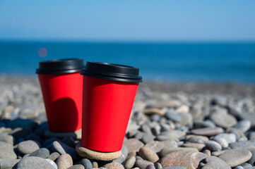 Close-up of couple of paper coffee cups on pebble beach. Blue sea on background. Hot take away drinks.