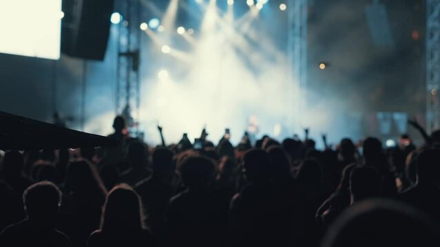 A blurry and illuminated dancing crowd. A crowd of fans standing near the stage applauding their favorite rock band. Concert in the city center. Young people with smartphones. Silhouettes of musicians