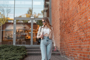 Fashionable pretty woman with trendy sunglasses in urban casual clothes with leather jacket, jeans and purse walks on the street near a brick building mall