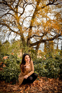 Chica mujer joven agachada frente un &aacute;rbol con hojas doradas en el suelo por la caida en oto&ntilde;o y con una hoja en la mano
