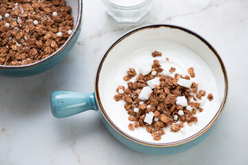 Turquoise bowl with greek yogurt and chocolate granola, elevated view on a light-beige marble background, horizontal shot