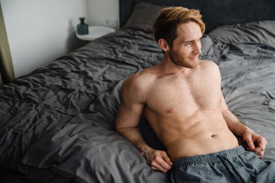 Young bearded shirtless man smiling and lying in bed at home