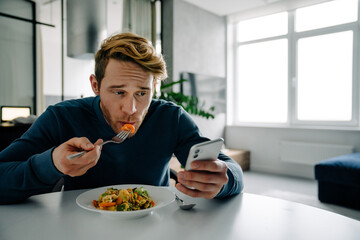 Young bearded white man eating dinner and using cellphone at home