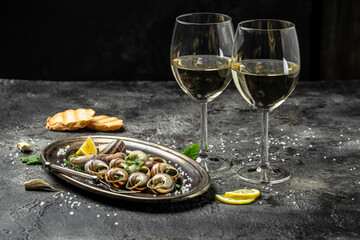 Delicious cooked sea escargo snails with herbs, butter, garlic on metal plate with forks. wine glass. gourmet food. Restaurant menu, Traditional French cuisine