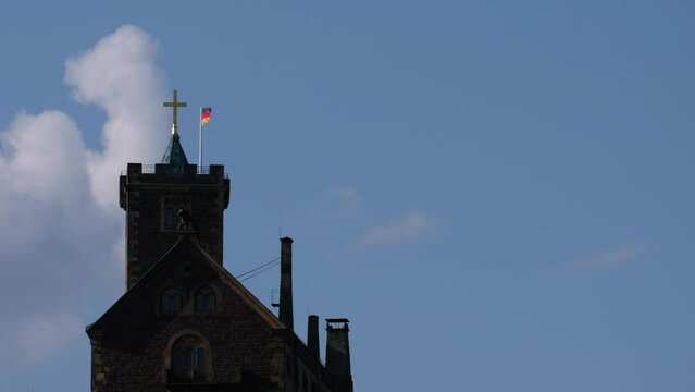 Waving german flag and a cross on the tower of the famous Wartburg Castle in Eisenach on a partly cloudy summer afternoon. Illustrious Unesco World Heritage palace in Thuringia, Germany.