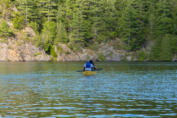 Couple in life vests paddling in a bright yellow kayak on a lake surrounded by rocks and thick coniferous forest, sunny summer day, selective focus. Water sports, recreation, adventure concept.