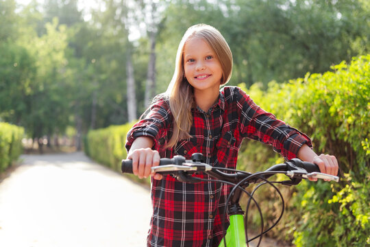 Smiling little girl stands with a bicycle and looking away in park. Kid riding bicycle outdoor