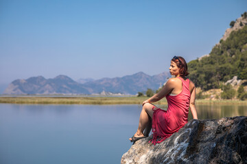 Naklejka na ściany i meble Beautiful girl in a red dress by the sea. Sunny day. Colorful sea landscape. Beautiful mountains. Girl by the sea. A girl in a dress. Summer day.