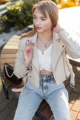 Fashionable young beautiful woman model with fashion streetwear with leather jacket and blue stylish jeans with handbag sits on the wooden bench near a cafe at sunset