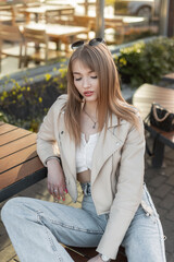 Fashionable beautiful female portrait of a pretty young hipster girl in a street fashion outfit with leather rock jacket and blue jeans sitting on a bench outside a cafe in the city