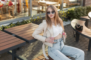 Fashionable beautiful stylish hipster girl model with round  sunglasses in a fashion leather rock jacket with a top and vintage blue jeans and a bag sitting on a wooden bench near a cafe at sunset