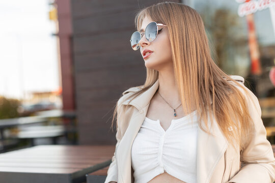 Street fashion portrait of trendy beautiful young woman hipster with stylish round sunglasses in rock fashionable leather and white top sits near a cafe in the city