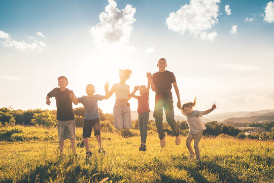 Six kids brothers and sisters teenagers and little kids jumping on a green grass meadow with evening sunset background light holding hands in hands and smiling. Happy and careless childhood concept