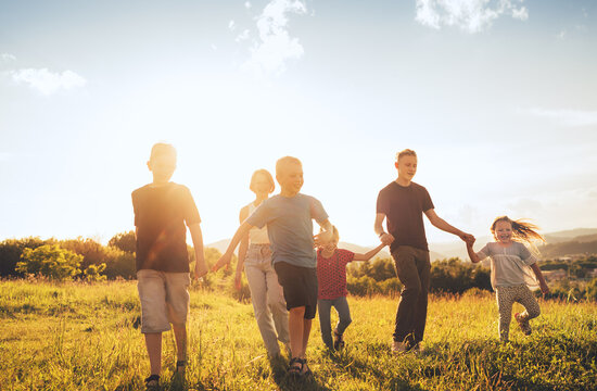 Six kids brothers and sisters teenagers and little kids walking on the green grass meadow with evening sunset background light holding hands in hands and smiling. Happy and careless childhood concept