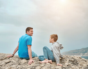 Fototapeta premium Happy family, father and son bonding, sitting on stone by the sea looking at view enjoying summer vacation. Togetherness Friendly concept 