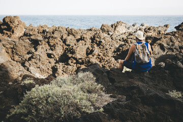 Fototapeta na wymiar Hiker with hat is sitting on volcanic rock, has backpack on his back, wearing blue shirt and looks out to sea, Atlantic Ocean