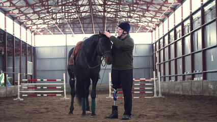 in special hangar, a disabled man jockey strokes a muzzle of a thoroughbred, black horse. man has...