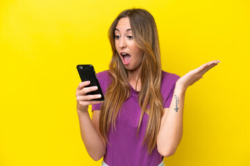 Naklejka premium Young caucasian woman isolated on yellow background looking at the camera while using the mobile with surprised expression