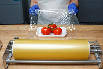 Unknown worker wraps in food transparent film tomatoes lying on white plastic tray.