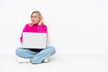 Young caucasian woman with laptop sitting on the floor listening to something by putting hand on the ear