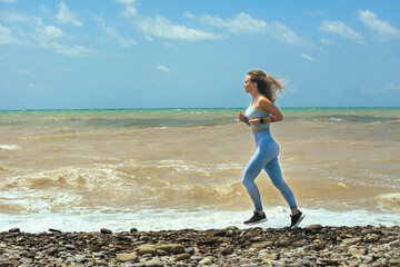 beautiful young female athlete in a blue tracksuit and beautiful wavy hair jogging along the sea on which there is a storm on a sunny day against a blue sky